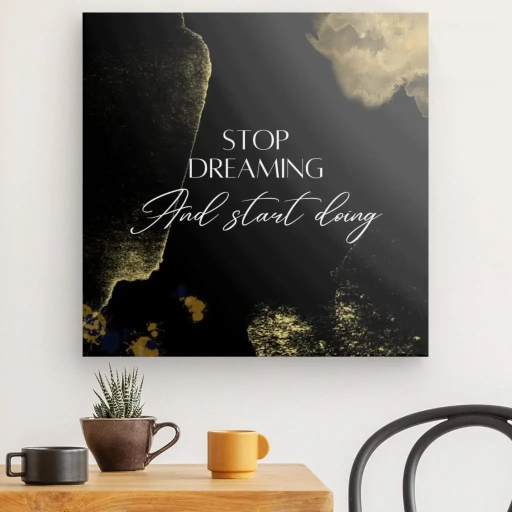 Une décoration murale carrée « Tableau 'Stop Dreaming' Design Noir Blanc et Doré » comportant le texte « Stop Dreaming And Start Doing » dans une police d'écriture blanche, montée au-dessus d'une table en bois contenant une petite plante et deux tasses. Une partie d'une chaise est visible à droite.