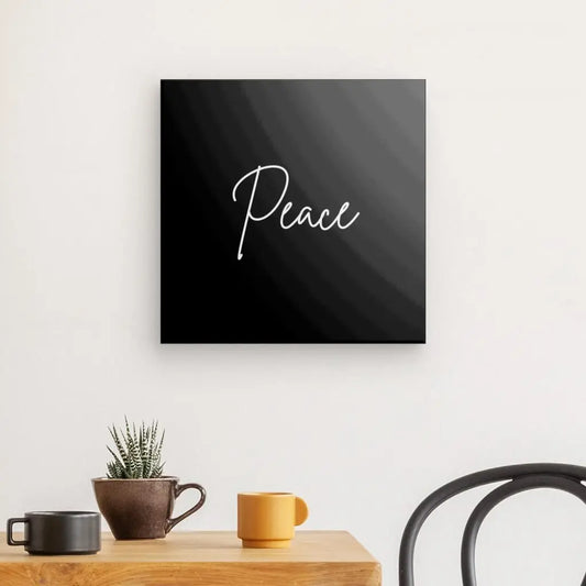 Une décoration murale avec le mot « Paix » en cursive est suspendue au-dessus d'une table en bois avec une petite plante en pot et deux tasses. Une partie d'une chaise noire est visible à droite, mettant en valeur ce Tableau "Peace" Minimaliste Noir et Blanc.