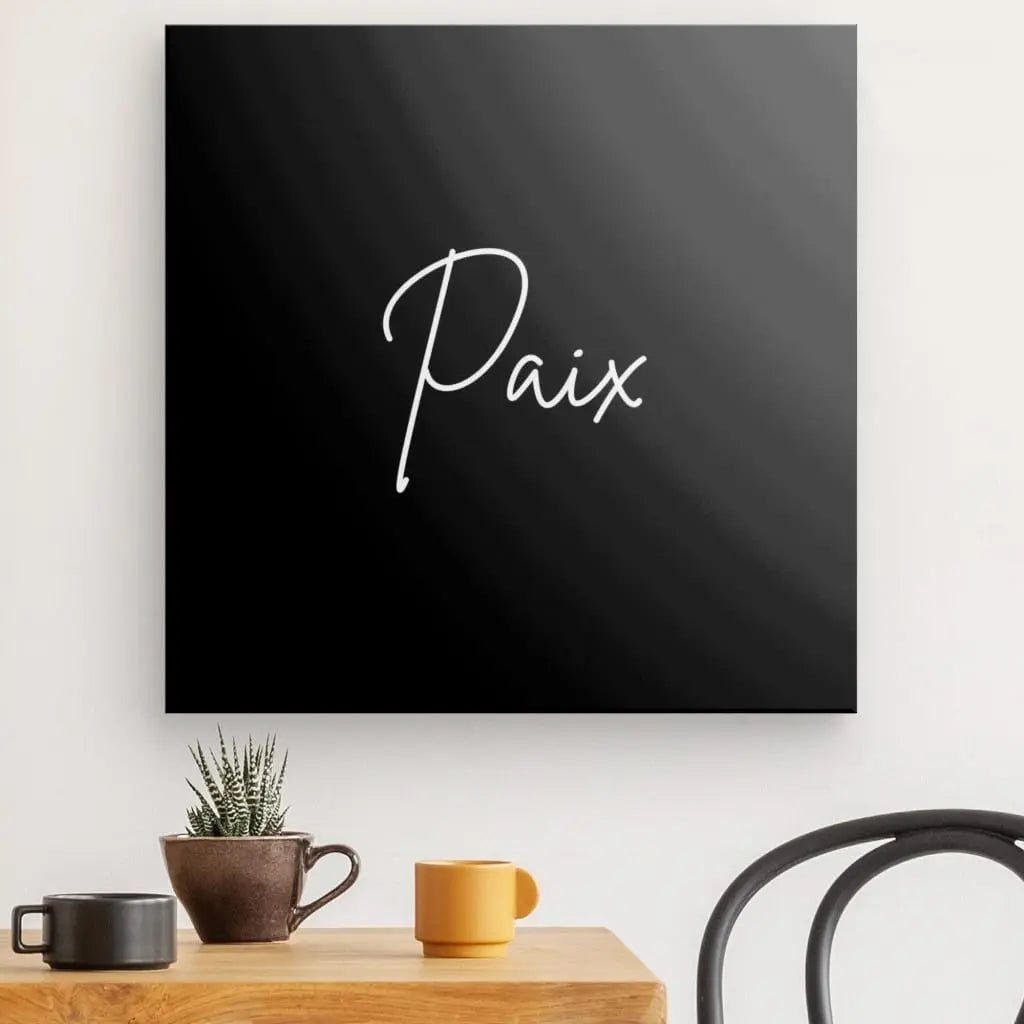 Une décoration murale carrée noire comportant le mot « Paix » en cursive blanche crée une atmosphère zen. En dessous, une table en bois contient une petite plante, une tasse noire et une tasse orange. Une chaise noire est partiellement visible à droite, complétant le Tableau Minimaliste "Paix" Noir et Blanc.