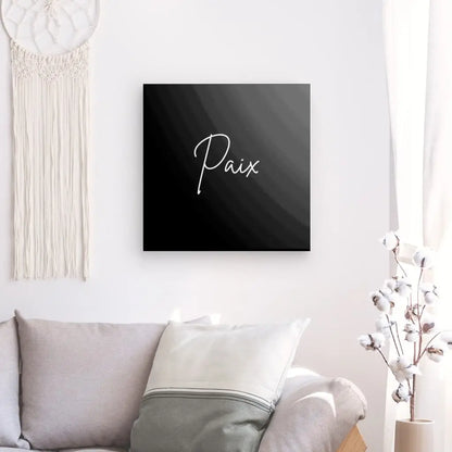 Tableau Minimaliste "Paix" Noir et Blanc avec le mot "Paix" en cursive blanche, suspendu au-dessus d'un canapé gris clair avec des coussins. Un rideau blanc et un tissage décoratif pendent à proximité, renforçant l'ambiance zen. Ce tableau minimaliste en noir et blanc ajoute une touche d'élégance sereine à votre espace de vie.