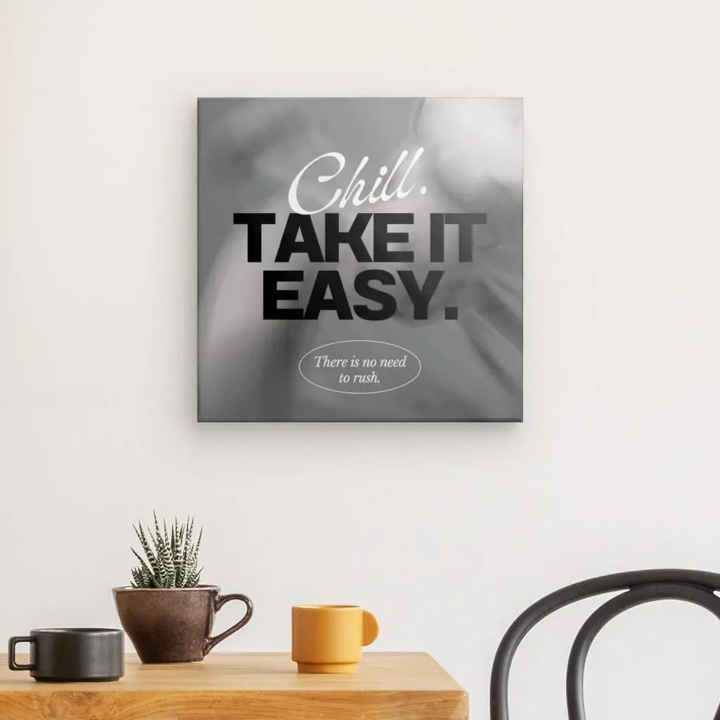 La déco murale Tableau "Take it easy" Noir et Blanc avec le texte "Chill. Take it easy. Il n'y a pas besoin de se précipiter." au dessus d'une petite table avec une plante, un mug et une bougie, à côté d'une chaise crée une ambiance apaisante parfaite pour toute décoration intérieure.