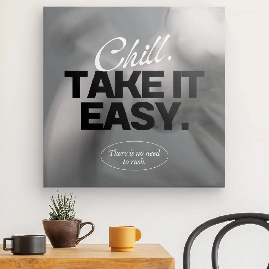 Une œuvre d'art mural de la collection Tableau "Take it easy" Noir et Blanc indique "Chill. TAKE IT EASY". ci-dessus "Il n'est pas nécessaire de se précipiter." Créant une ambiance apaisante, une petite plante et deux tasses reposent sur une table avec une chaise à côté, rehaussant la décoration intérieure en tableau noir et blanc.