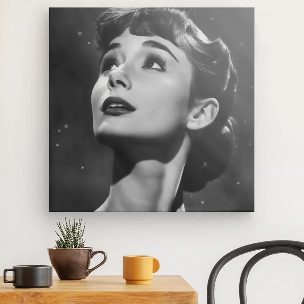 Le portrait encadré en noir et blanc d'une femme est suspendu au-dessus d'une table en bois avec une petite plante en pot, une tasse noire et une tasse orange. Une chaise noire est partiellement visible à côté de la table, ajoutant une touche d'élégance qui rappelle le Tableau Audrey Hepburn Noir et Blanc.