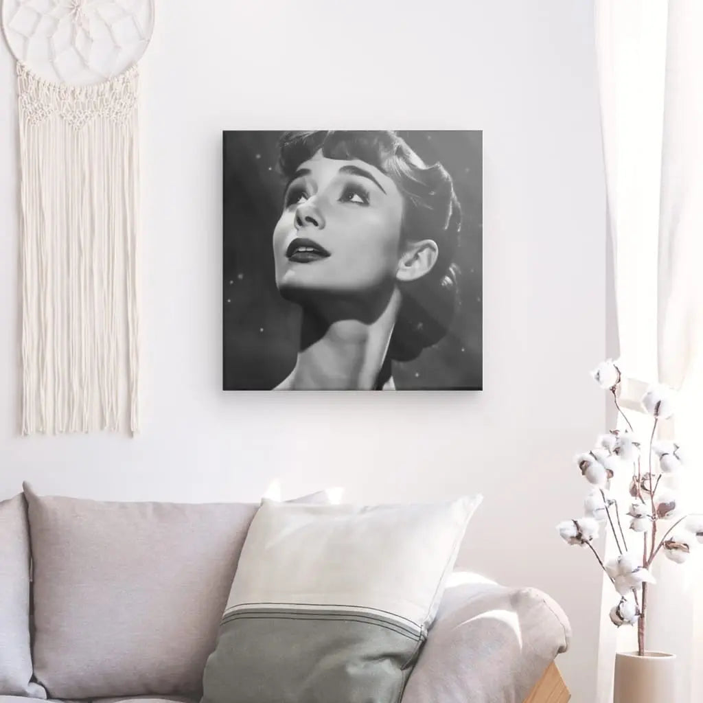 Un Tableau Audrey Hepburn Noir et Blanc est encadré et accroché sur un mur blanc au-dessus d'un canapé gris clair avec un coussin blanc. Une tenture murale en macramé se trouve à gauche et un rideau blanc à droite, créant un tableau élégant.