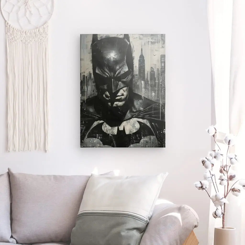 Un "Tableau Batman Peinture Noir et Blanc" encadré est accroché sur un mur blanc au-dessus d'un canapé gris avec des oreillers blancs et gris. À côté se trouve un tableau d'Audrey Hepburn, présentant un élégant décor intérieur noir et blanc, complété par une tenture murale en macramé blanc et des tiges de coton dans un vase en verre.