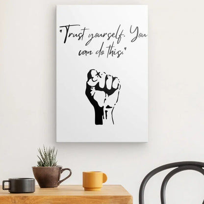 Le Tableau Citation Confiance Poing Noir et Blanc de haute qualité présente une citation motivante "Trust yourself. You can do this." au-dessus d'une illustration d'un poing levé. En dessous, il y a une table avec une plante en pot, une tasse jaune et un bol orange.