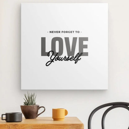 Une citation encadrée « Tableau Love Yourself Noir et Blanc » avec le texte « NEVER FORGET TO LOVE Yourself » est placée au-dessus d'une table en bois contenant un petit cactus en pot, une tasse jaune et une tasse orange.