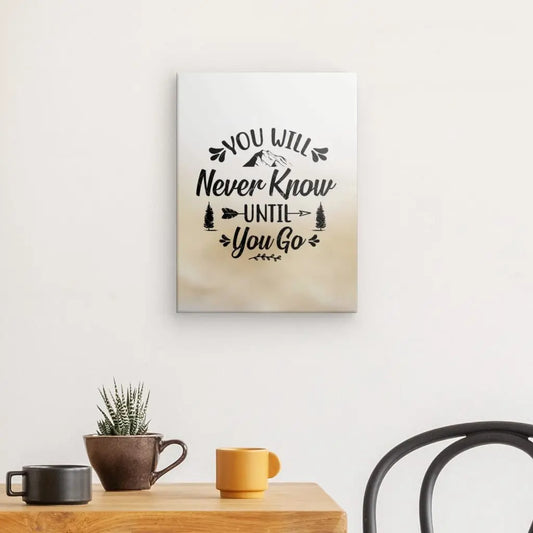 Une toile murale intitulée "Tableau Citation 'Never Know' Noir Blanc Beige" avec la citation "Vous ne le saurez jamais jusqu'à ce que vous partiez" est suspendue au-dessus d'une table en bois qui contient une plante en pot et deux tasses, une noire et une jaune. Cette citation de tableau ajoute un esprit d'aventure à la déco moderne de la pièce. Une chaise est partiellement visible à droite.