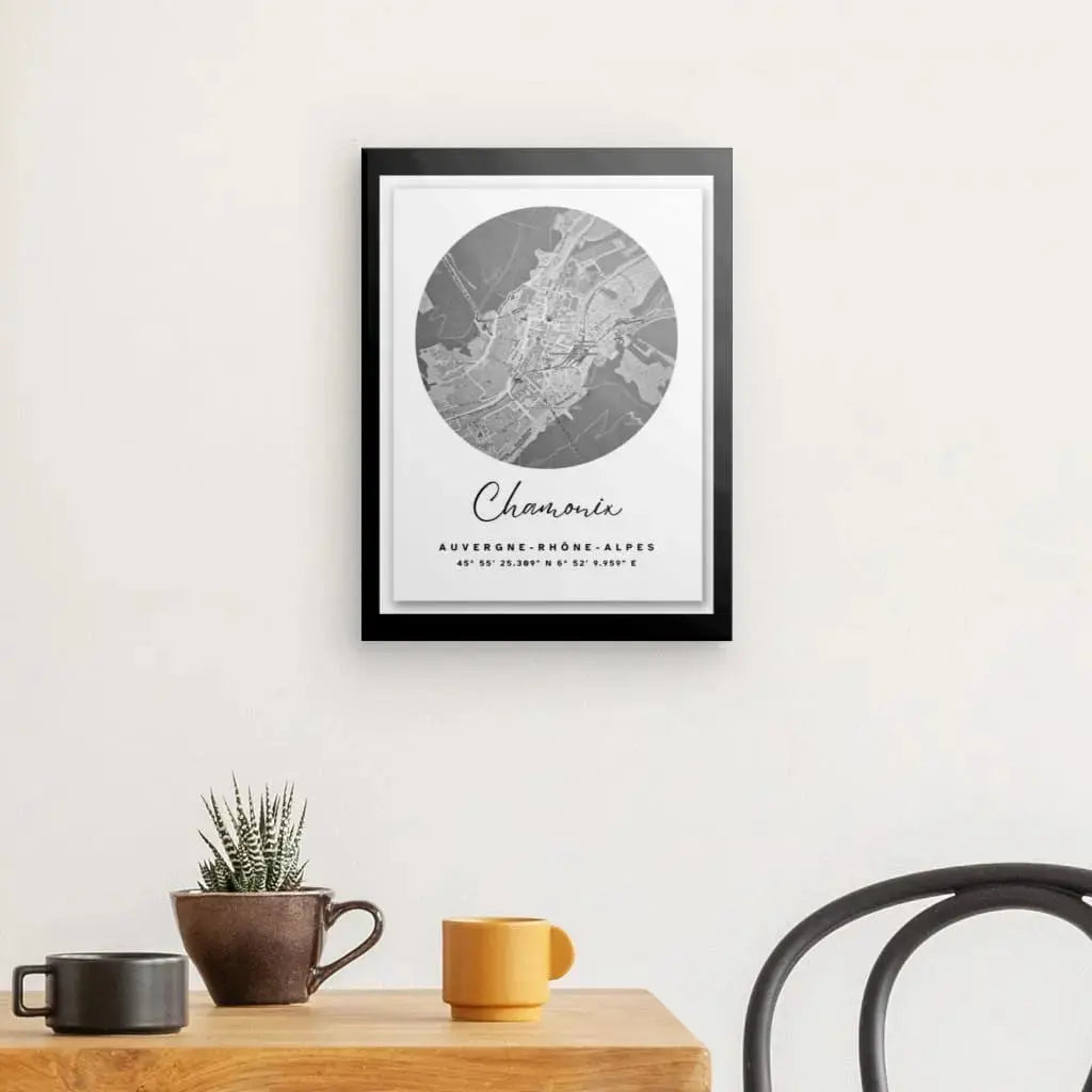 Un Tableau Carte Ville Chamonix Noir et Blanc encadré est accroché sur un mur blanc au-dessus d'un bureau en bois avec une petite plante, une tasse grise et une tasse orange. Une chaise au dossier courbé est partiellement visible à droite, créant un espace de travail harmonieux orné de cette belle œuvre d'art.