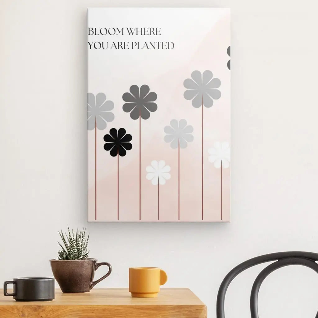 Un magnifique Tableau Citation Floraison Fleur Noir Blanc Rose avec des fleurs stylisées grises, noires et blanches et la citation « Fleurissez là où vous êtes planté » est suspendu au-dessus d'une table en bois ornée d'une petite plante en pot et de trois tasses à café.