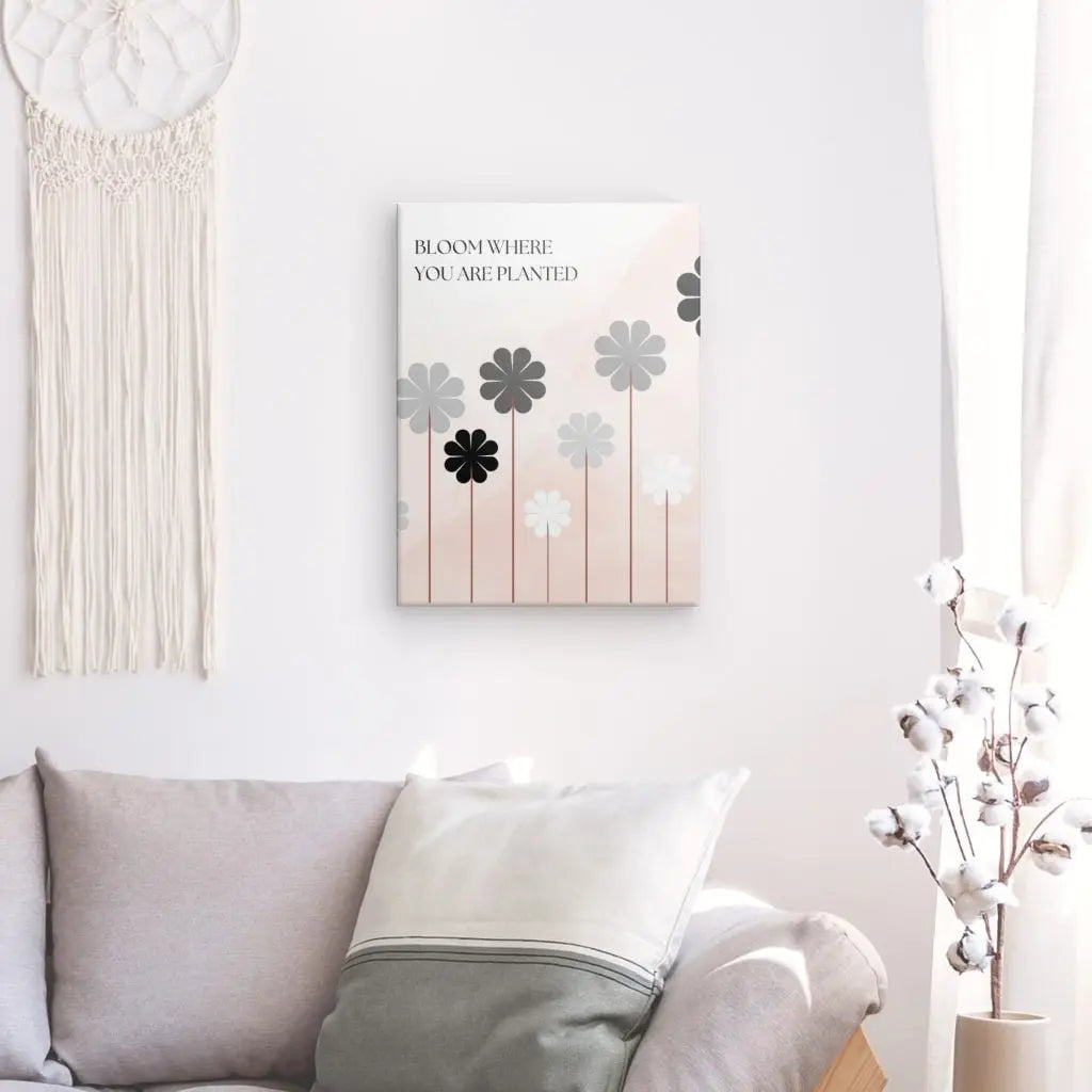 Une décoration murale avec le texte « Fleurissez où vous êtes planté » (Tableau Citation Floraison Fleur Noir Blanc Rose) est suspendue au-dessus d'un canapé doté de coussins. Une tenture murale en macramé est à gauche, et un vase avec des tiges de coton est à droite, complétant la charmante décoration intérieure.