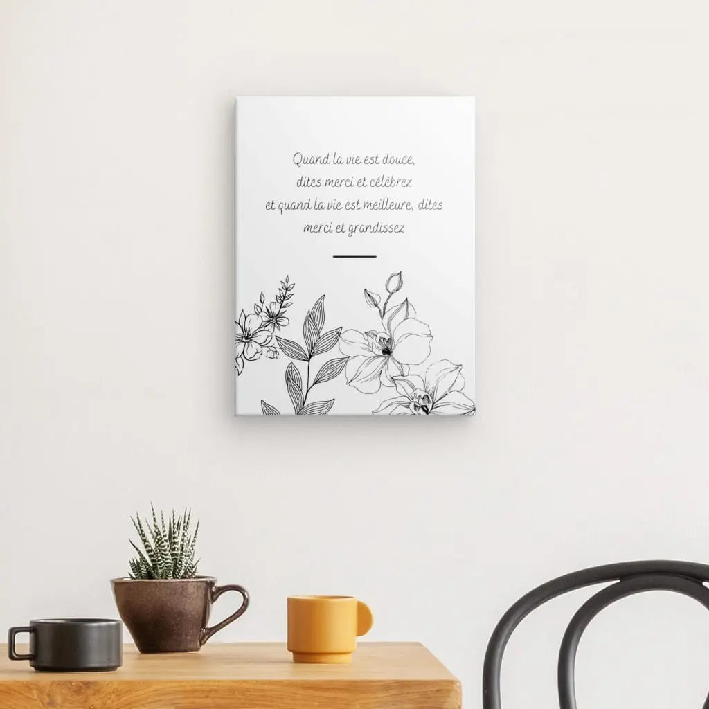 Une œuvre d'art mural avec un texte en français et un Tableau Citation Fleurs et Gratitude Inversé est suspendue au-dessus d'une table en bois sur laquelle se trouvent un petit cactus, une tasse marron et une boisson orange. La décoration intérieure élégante comprend une chaise noire à proximité.