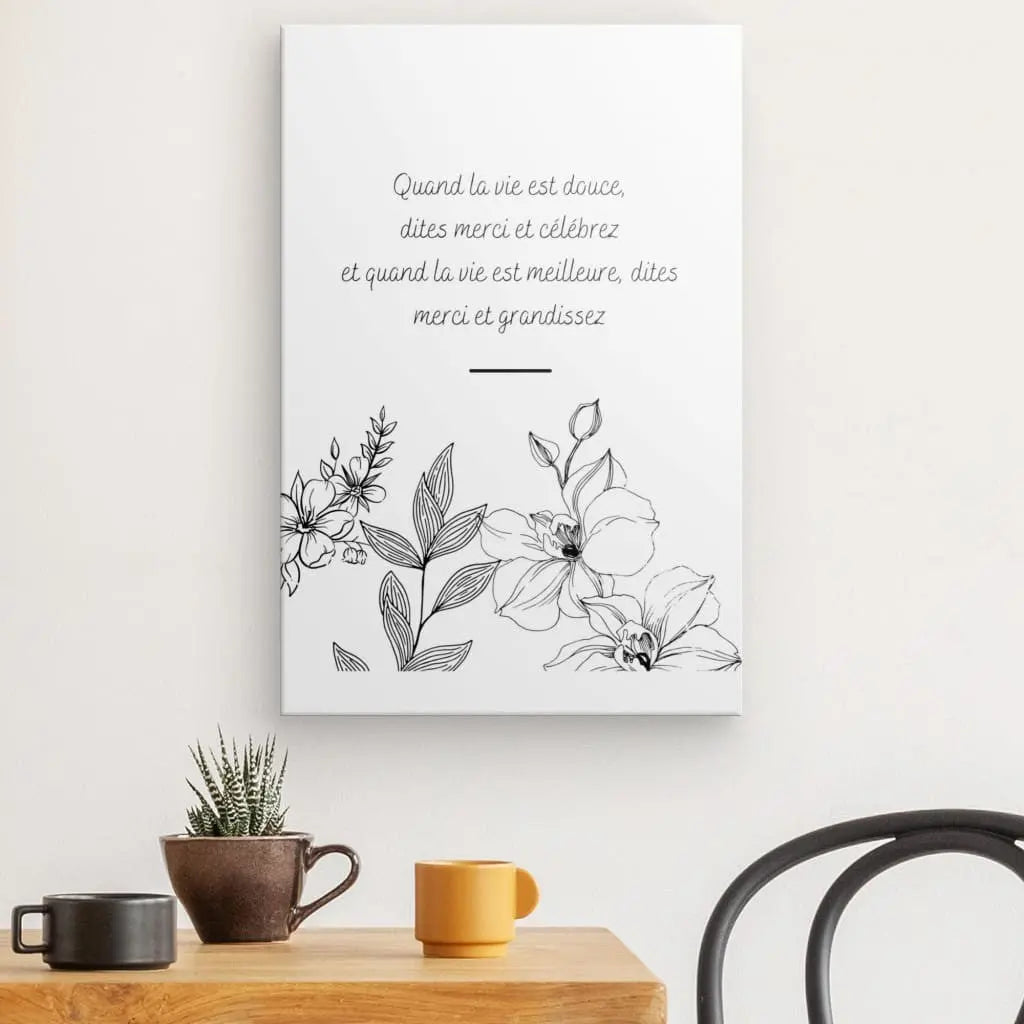 Un Tableau Citation Fleurs et Gratitude Inversé comportant une citation française et entouré de croquis floraux est suspendu au-dessus d'une petite table en bois avec un cactus en pot, une tasse jaune, une tasse orange et une chaise noire. Cette décoration intérieure élégante ajoute du charme à la pièce.