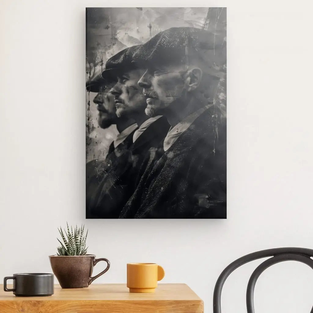 Le Tableau Peaky Blinders Noir et Blanc représente trois hommes de profil portant des casquettes plates et des vêtements sombres, rappelant Peaky Blinders. Ce tableau noir et blanc est accroché au-dessus d'une table en bois avec une petite plante et trois tasses, à côté d'une chaise noire, parfait pour une décoration intérieure sophistiquée.