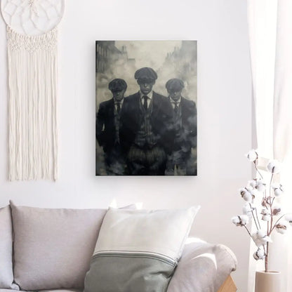 Un Tableau Peinture Gang Peaky Blinders Monochrome est accroché à un mur au-dessus d'un canapé beige avec des oreillers. Une tenture murale en macramé est à gauche, et un vase avec des branches de coton complète la décoration intérieure à droite.