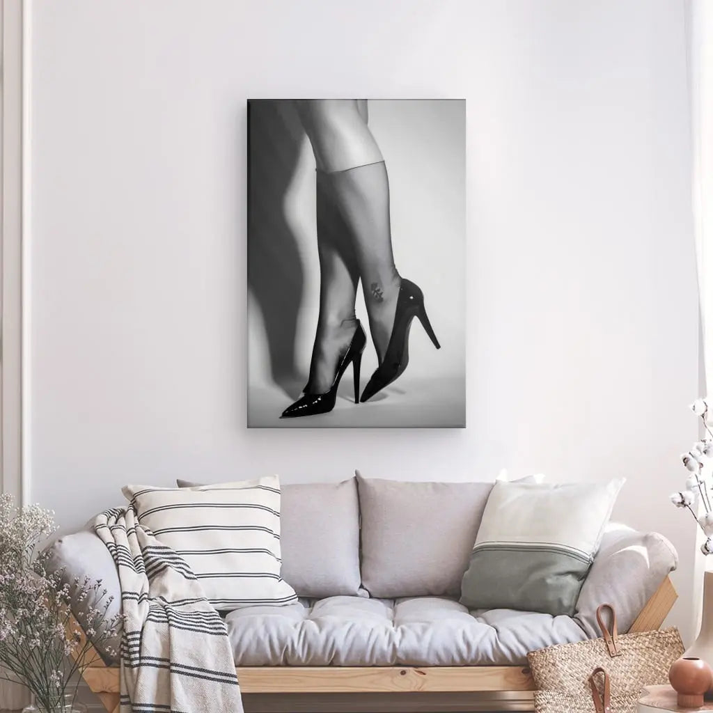 Un salon comprend un canapé gris clair avec des oreillers et une couverture rayée, sous une photo en noir et blanc intitulée "Tableau Résille Jambes Femme Escarpins Noir et Blanc", mettant en valeur les jambes en bas transparents et talons hauts. Un panier, des plantes décoratives et un tableau décoration intérieure complètent le cadre cosy.