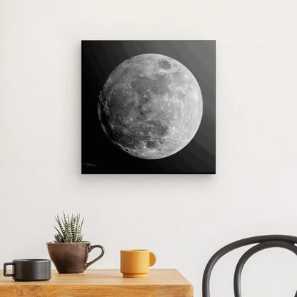 Une photographie en noir et blanc intitulée "Tableau Lune Gros Plan Noir et Blanc" est accrochée sur un mur blanc au-dessus d'une table en bois clair avec une petite plante en pot et deux tasses, une orange et une marron foncé. Cette œuvre d'art captivante est complétée par une chaise au dossier incurvé à droite.