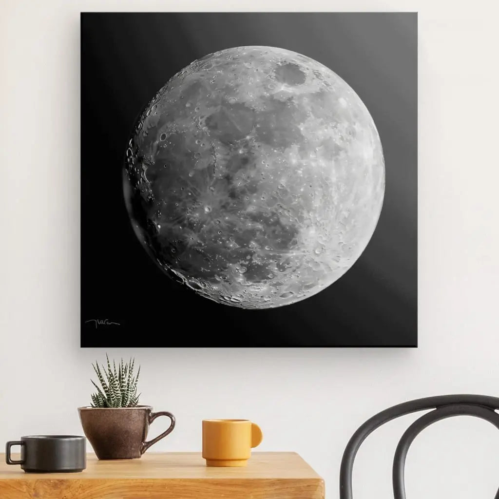 Un Tableau Lune Gros Plan Noir et Blanc encadré de la surface lunaire de la lune est accroché sur un mur blanc au-dessus d'une table en bois avec une plante en pot et deux tasses. Une chaise noire est partiellement visible à droite, ajoutant à la composition sereine de cette œuvre d'art.