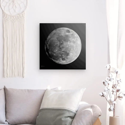 Un Tableau Lune Gros Plan Noir et Blanc est accroché sur un mur blanc au-dessus d'un canapé de couleur claire avec un oreiller blanc. Cette œuvre d'art captivante est complétée par une tenture murale en macramé et un vase à fleurs blanches.