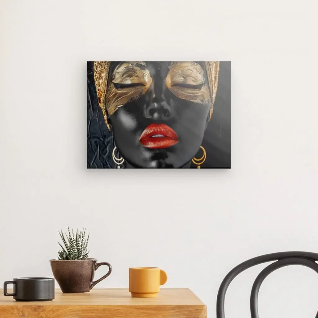 Une photo encadrée, **Tableau Femme Portrait Moderne Dorée Rouge Noir**, représentant une femme avec de la peinture noire, des masques pour les yeux dorés et du rouge à lèvres, est accrochée sur un mur blanc au-dessus d'une petite table avec des plantes en pot et une tasse jaune et marron. décoration intérieure classique. Une chaise noire est partiellement visible.