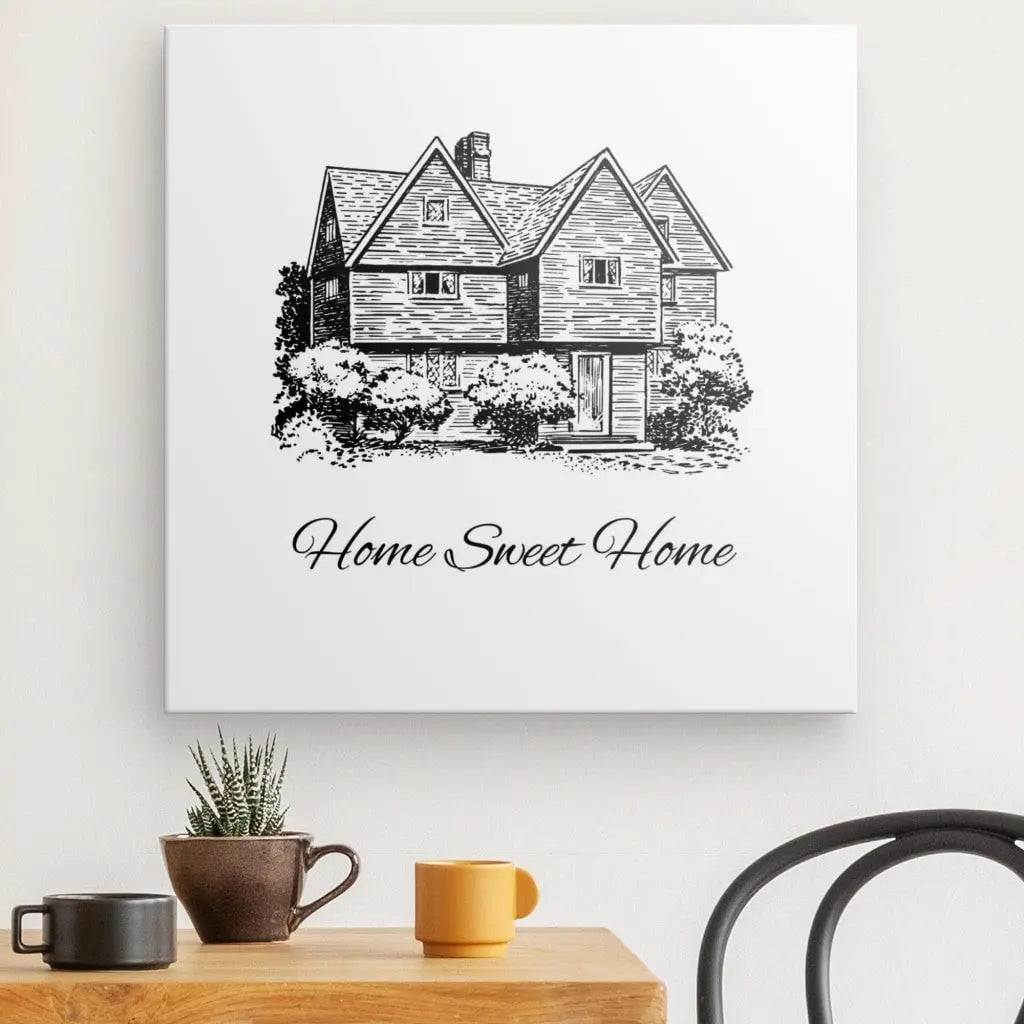 Une œuvre d'art encadrée intitulée "Tableau Home Sweet Home Blanc et Noir", représentant une charmante maison et le texte "Home Sweet Home", est exposée sur un mur blanc au-dessus d'une table en bois décorée d'une petite plante et de deux tasses. Cette pièce de décoration intérieure ajoute du charme à n'importe quelle pièce.