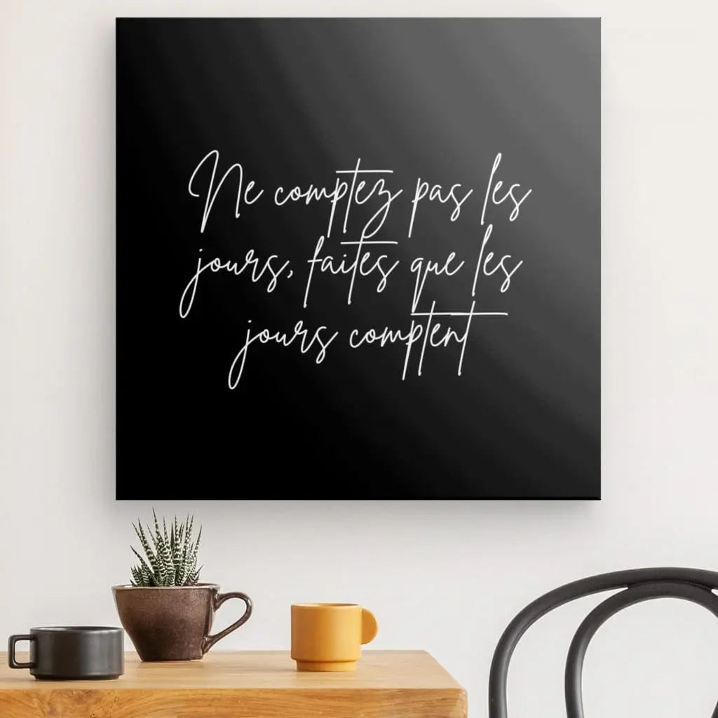 La citation Tableau « Ne comptez pas les jours » Noir et Blanc, avec la citation française « Ne comptez pas les jours, faites que les jours comptent » en cursive blanche, ajoute une touche de citation inspirante. En dessous, une table ornée d'une petite plante, d'une tasse marron et d'une tasse orange rehausse la décoration intérieure élégante.