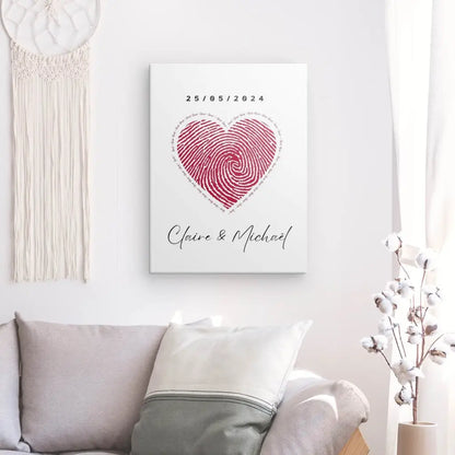 Une toile encadrée sur un mur pâle affiche un motif d'empreinte digitale rouge en forme de cœur avec la date « 25/05/2024 » et les noms « Claire & Michael » en dessous, créant un élégant Tableau Couple Personnalisé Coeur qui rehausse la décoration intérieure minimaliste.