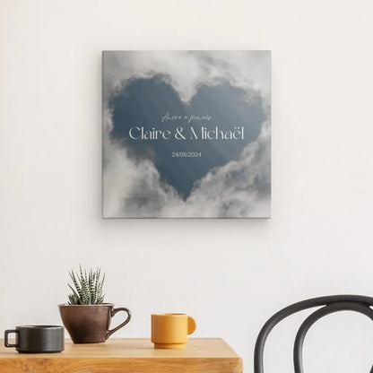 Art mural d'un coeur en forme de nuage avec texte "Claire & Michael" et date "24/05/2024". Ci-dessous se trouve une table avec une plante en pot, une tasse jaune et une tasse sombre, à côté d'une chaise noire. Ce Tableau Personnalisé Amoureux Coeur Nuages est une impression haute définition sur toile de qualité supérieure.