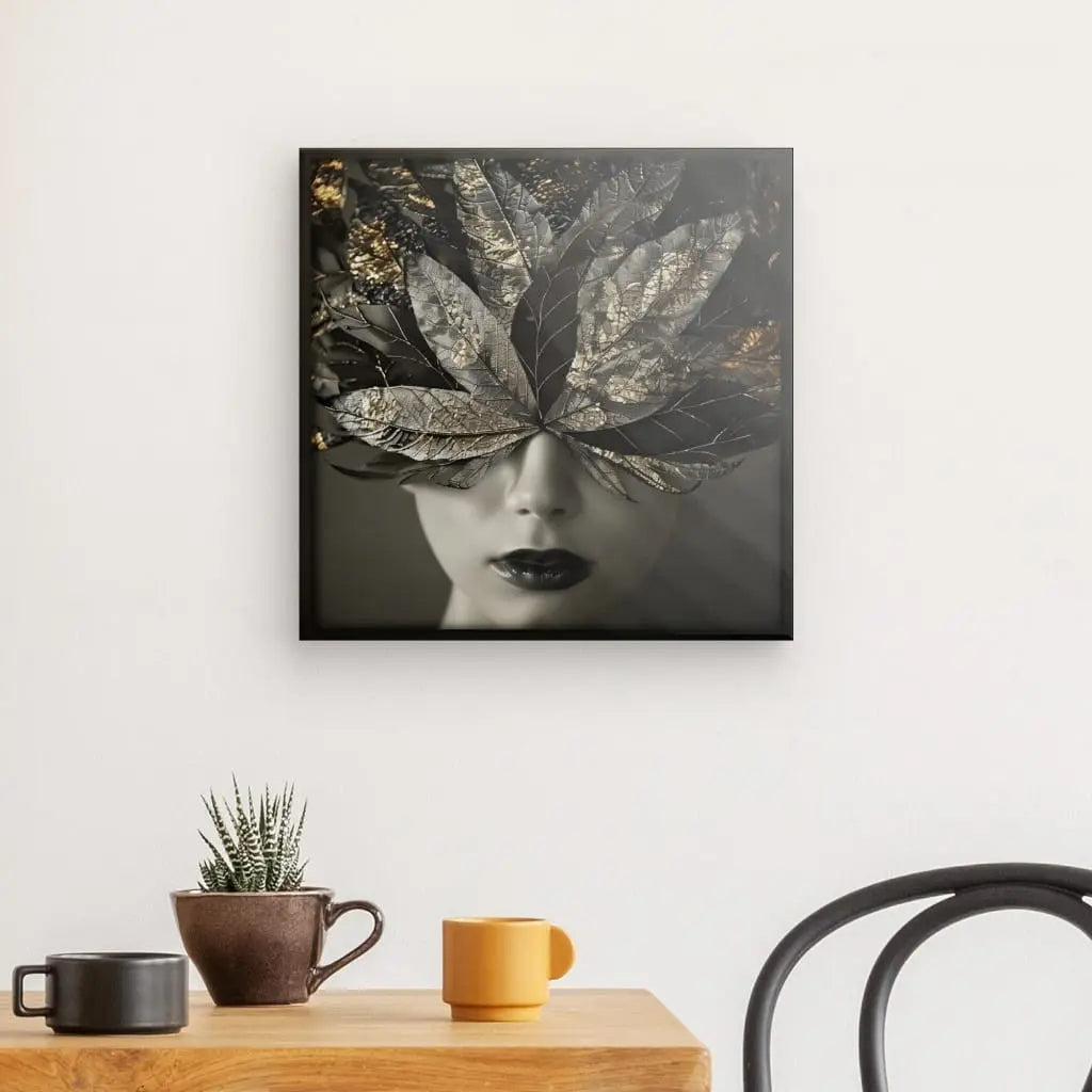 Un mur présente un saisissant « Tableau Visage Femme Plumes Dorées Noir et Blanc » représentant un visage de femme partiellement caché par des feuilles. En dessous, une table en bois avec une petite plante en pot, une tasse jaune, une tasse marron et une chaise noire rehaussent la décoration contemporaine.