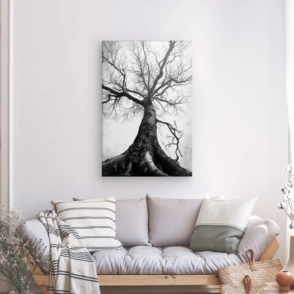 Le "Tableau Arbre Centenaire Noir et Blanc" est accroché au-dessus d'un canapé gris clair orné de coussins rayés et unis. Un panier tressé et une plante en pot sont placés à proximité, contribuant à la décoration élégante.