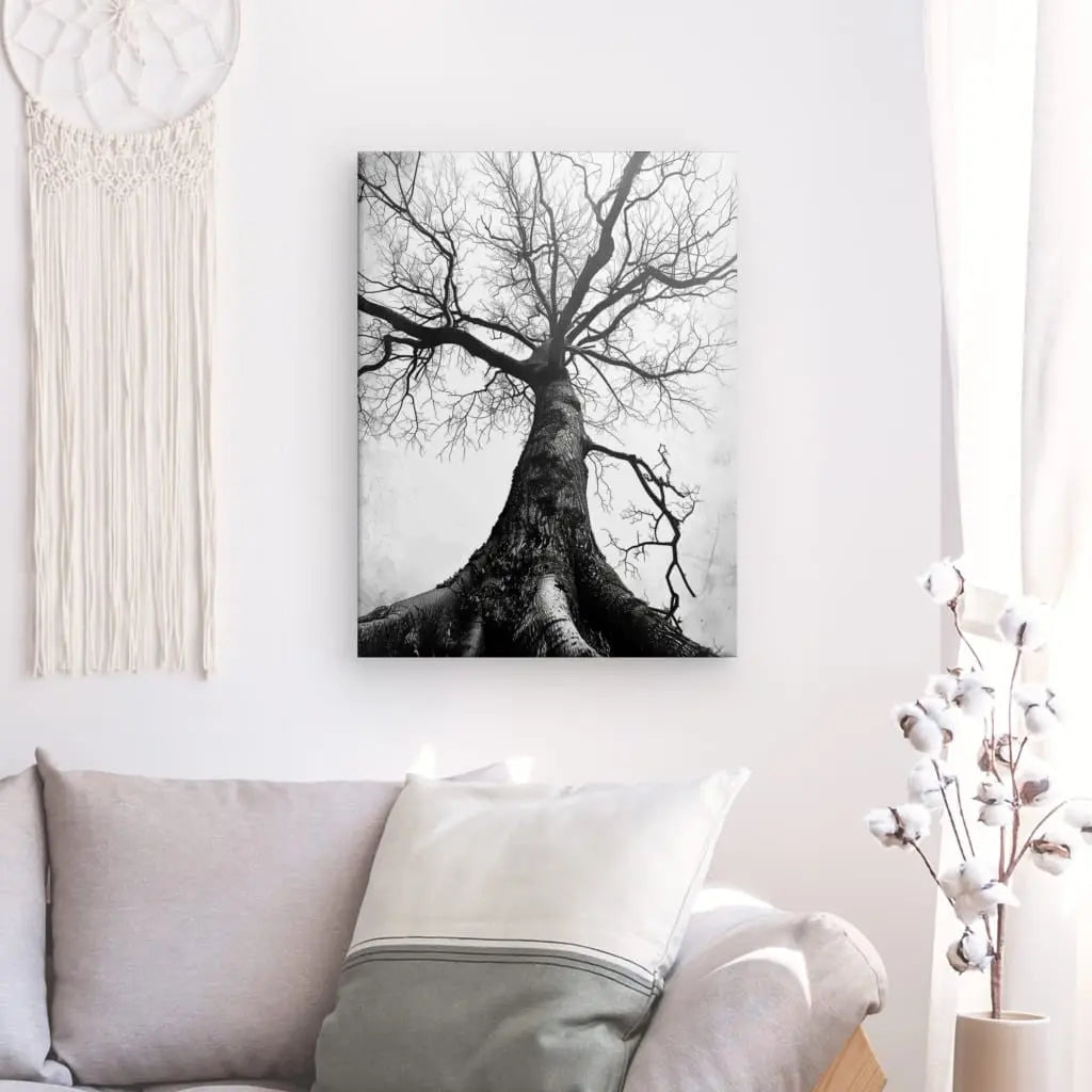 Le Tableau Arbre Centenaire Noir et Blanc d'un arbre est encadré sur un mur blanc au-dessus d'un canapé gris avec des oreillers blancs et gris. Une tenture murale en macramé est à gauche, ajoutant à la décoration élégante, avec un vase de fleurs en coton à droite.