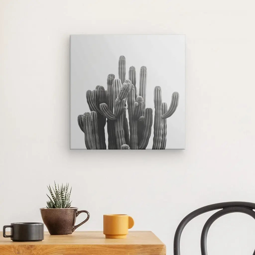 Un Tableau Carré Cactus Noir et Blanc est suspendu au-dessus d'une table en bois sur laquelle se trouve une petite plante en pot et deux tasses, une brune et une jaune. Une chaise noire est partiellement visible à droite, complétant cette décoration minimaliste.