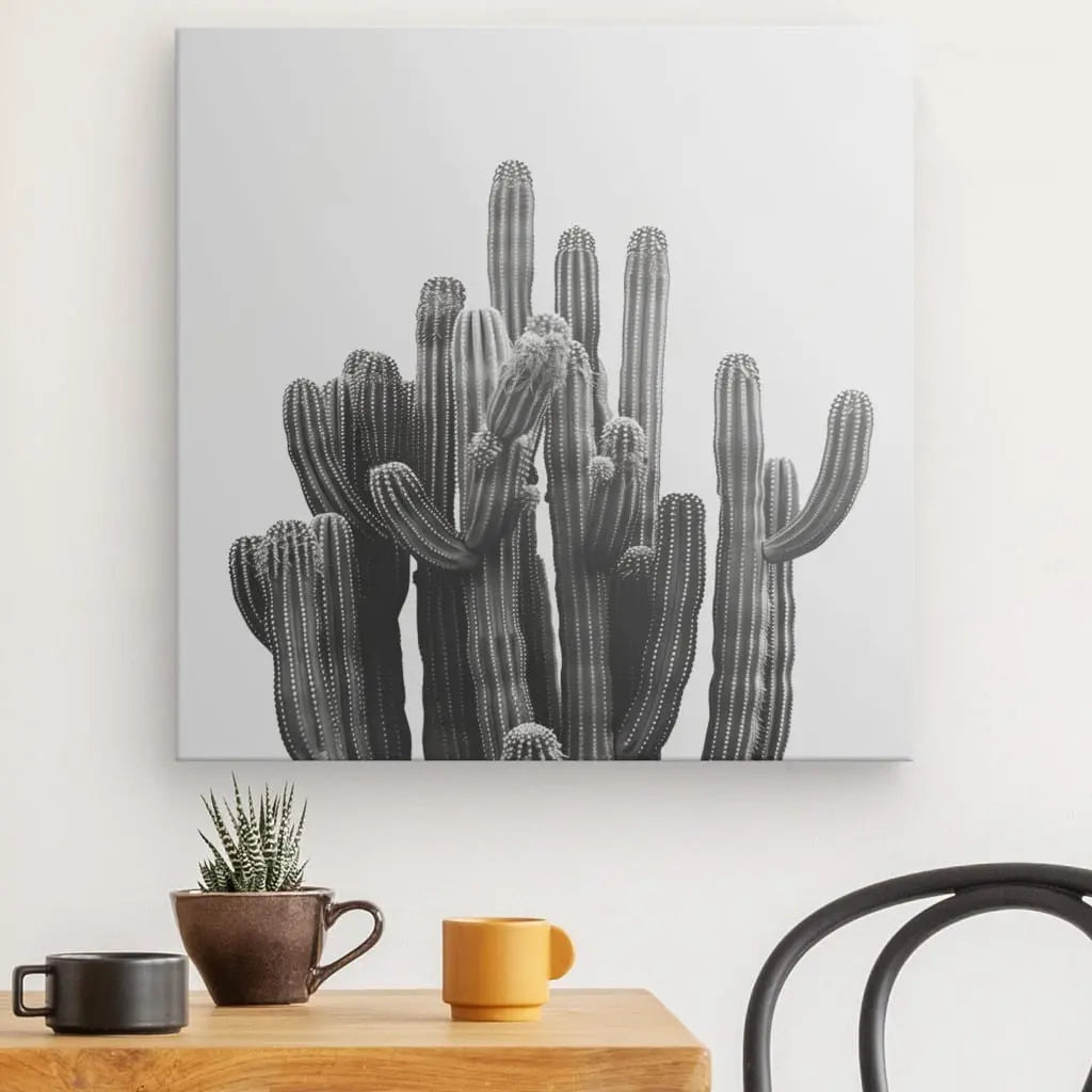 Un Tableau Carré Cactus Noir et Blanc est accroché au mur au-dessus d'une table en bois ornée d'un petit cactus en pot, d'une tasse jaune, d'une tasse marron et d'une chaise noire, créant un superbe tableau noir et blanc qui illustre une décoration minimaliste.