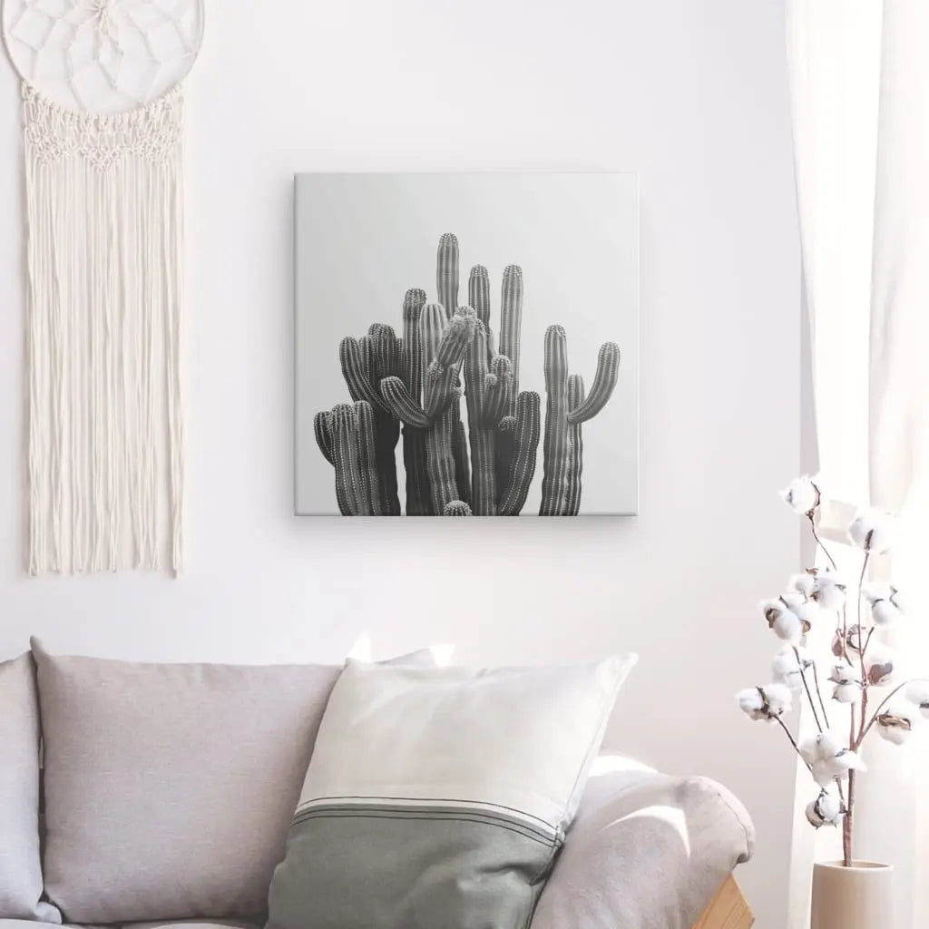 Un salon comprend un canapé gris avec un oreiller blanc, un Tableau Carré Cactus Noir et Blanc au mur, une tenture murale en macramé et des tiges de coton blanches dans un vase : parfait pour ceux qui apprécient la décoration minimaliste.