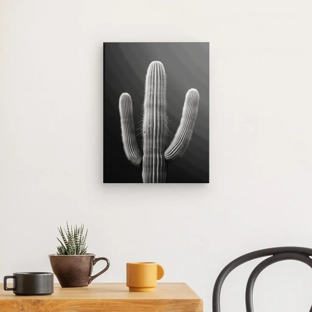 Une photo en noir et blanc d'un cactus, le "Tableau Cactus Désert Monochrome Fond Noir", est accrochée sur un mur blanc au-dessus d'une table en bois avec une petite plante de cactus, une tasse marron et une tasse jaune. Une chaise noire est partiellement visible à droite.