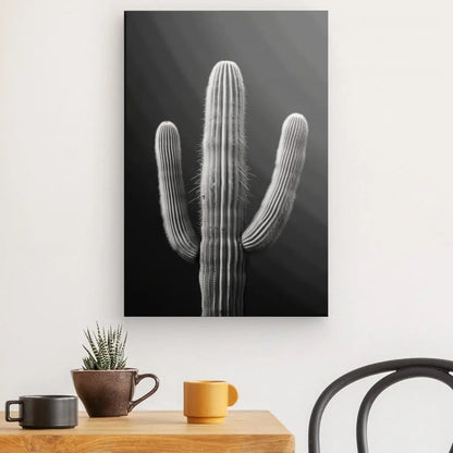 Un tableau cactus désert monochrome fond noir est accroché sur un mur blanc au-dessus d'une table en bois avec deux tasses et une petite plante en pot, à côté d'une chaise noire. Cette décoration contemporaine minimaliste respire la simplicité et l'élégance.