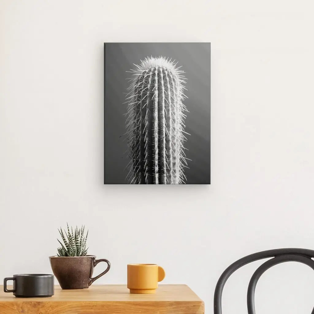 Une photographie en noir et blanc du « Tableau Cactus Gros Plan Minimaliste Monochrome » est affichée sur un mur blanc au-dessus d'une table en bois, qui contient un petit cactus en pot, une tasse marron et une tasse jaune. Une partie d'une chaise noire est visible à droite, créant une ambiance d'art minimaliste dans un décor contemporain.