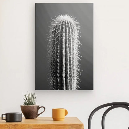 Le Tableau Cactus Gros Plan Minimaliste Monochrome est accroché sur un mur blanc au-dessus d'une table en bois ornée de petites plantes, d'un mug jaune et d'un mug noir. La scène résume l’essence de l’art minimaliste dans un décor contemporain.