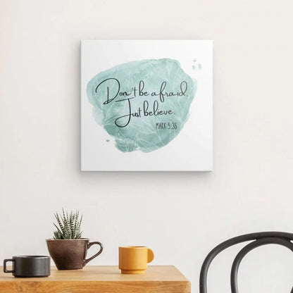 Le tableau "N'ayez pas peur, croyez juste" Noir Blanc Touche Verte est accroché au mur, offrant une citation inspirante. En dessous, une table en bois accueille une petite plante en pot, un mug noir et un mug orange, créant une décoration contemporaine harmonieuse.