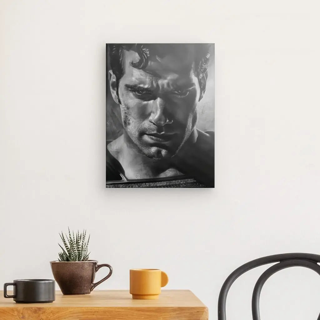 Un portrait en noir et blanc d'un homme, intitulé "Tableau Portrait Gros Plan Superman Noir et Blanc", est accroché sur un mur blanc au-dessus d'une table en bois ornée d'une plante en pot, d'une tasse marron et d'une tasse jaune, créant ainsi un noir et blanc. tableau qui met en valeur la décoration intérieure. Une chaise noire est partiellement visible à droite.