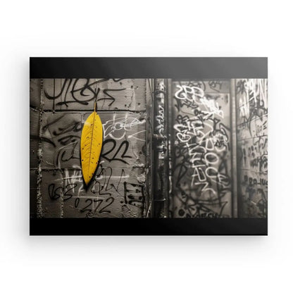 Un Tableau Underground Street Noir Blanc Feuille Jaune est monté sur un mur de béton recouvert de graffitis dans une ruelle urbaine faiblement éclairée, ajoutant une touche de décoration contemporaine au street art vibrant qui décore l'espace.