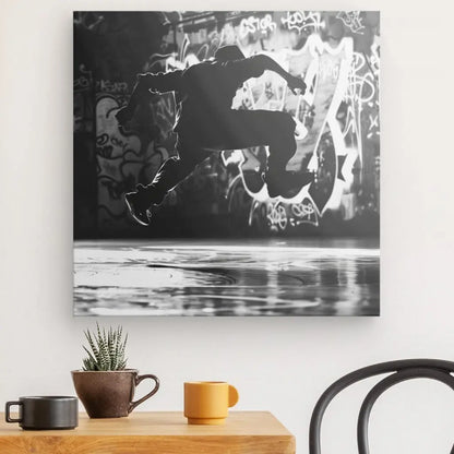 Une photo monochrome Tableau Breakdance Danseur Hip-Hop sur un mur, représentant un danseur hip-hop en plein saut devant une zone couverte de graffitis. Une table en bois avec une plante en pot, une tasse marron et une tasse jaune est au premier plan, créant un tableau d'art contemporain.