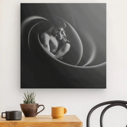 Le Tableau Couple Etreinte Douce Noir et Blanc, représentant un homme torse nu embrassant une femme, est exposé sur un mur au-dessus d'une table avec une plante et deux mugs colorés, ajoutant une touche de décoration romantique à votre espace.