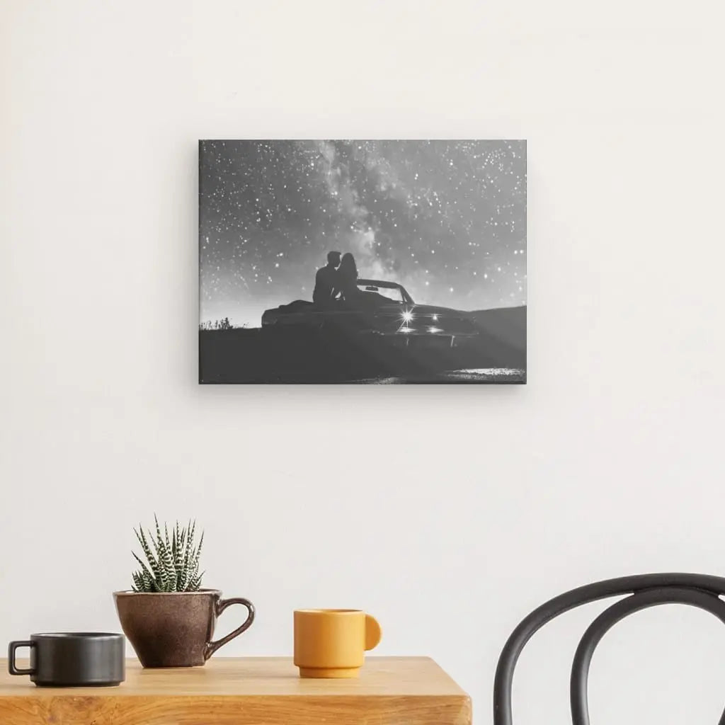 Le Tableau Couple Nuit étoilée Voiture Noir et Blanc, représentant une photographie en noir et blanc de deux personnes assises dans une voiture ancienne sous un ciel étoilé, est élégamment exposé sur un mur blanc au-dessus d'une table en bois ornée d'une petite plante, d'un mug noir, et une tasse jaune. Cette pièce captivante ajoute du charme à votre décoration intérieure.