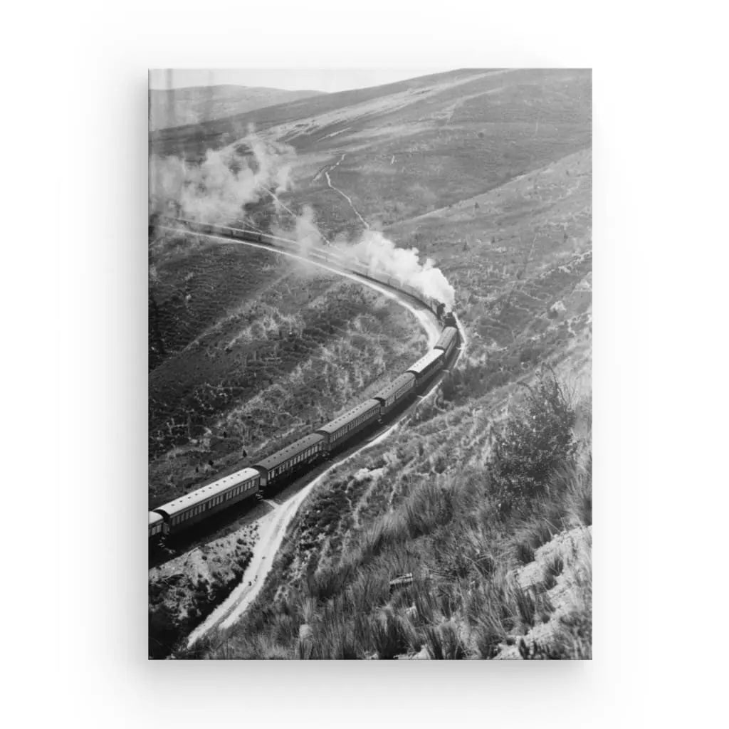 Le Tableau Train à Vapeur Paysage Noir et Blanc capture un train à vapeur voyageant à travers un paysage montagneux, émettant de la fumée lorsqu'il se déplace le long des voies courbes. Une végétation clairsemée recouvre les flancs des collines, créant un cadre pittoresque.