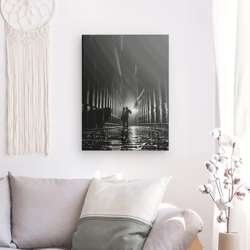 Un "Tableau Photo Noir et Blanc Couple Sous La Pluie" est accroché sur un mur blanc au dessus d'un canapé beige avec des coussins blancs et beiges. Des branches de coton dans un vase blanc rehaussent délicatement l'ambiance.