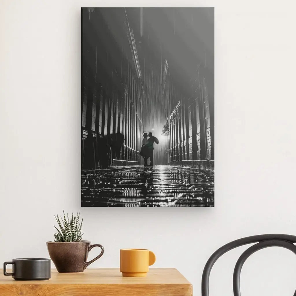 Un tableau photo noir et blanc accroché au mur, intitulé « Tableau Photo Noir et Blanc Couple Sous La Pluie », représente un couple marchant ensemble sous la pluie dans une ruelle étroite et humide, la nuit. L'image est accrochée au-dessus d'une table en bois ornée d'une petite plante en pot et de trois tasses, créant une scène évocatrice d'art photographique.