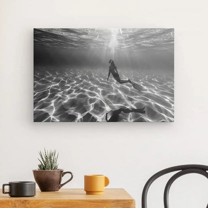 Accrochée sur un mur blanc au-dessus d'une table en bois avec deux tasses et une petite plante en pot, se trouve la photo en noir et blanc d'un nageur sous-marin, « Tableau Plongeuse Fond Sous Marin Noir et Blanc ». Une chaise noire est partiellement visible à côté de la table.