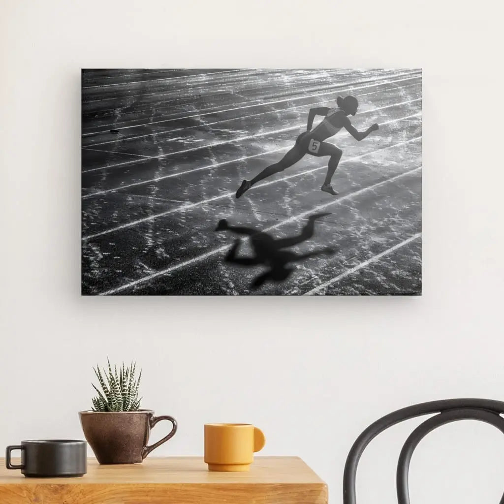 Un Tableau Athlétisme Coureur Noir et Blanc représentant un athlète en pleine course orne le mur au-dessus d'une table en bois avec deux tasses, une petite plante et une chaise à proximité.