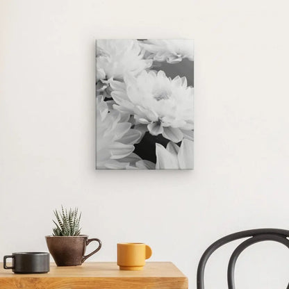 Une photographie en noir et blanc du Tableau Fleurs de Lotus Blanc Gros Plan est accrochée sur un mur blanc au-dessus d'une table en bois avec une petite plante en pot et deux tasses à café, se fondant parfaitement dans le décor intérieur chic. Une partie d'une chaise noire est visible à droite.